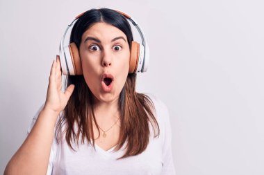 Genç, güzel esmer kadın müzik dinliyor. Kulaklıkları beyaz arka planda kullanıyor. Şaşırmak için ağzı açık ve korkmuş.