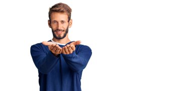 Sarışın, sakallı, sıradan bir süveter giyen, elleri avuçlarında gülümseyen ve el hareketi yapan yakışıklı bir adam. Bekleme ve koruma 