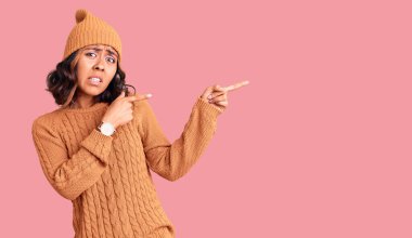 Yün kazak ve kış şapkası giyen genç, güzel, melez bir kadın endişeli ve endişeli bir ifadeyle iki elini de işaret ediyor. 