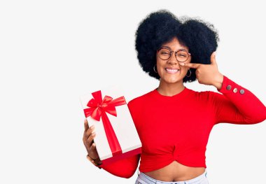 Genç Afro-Amerikalı kız elinde hediyeyle el ve burun buruna parmakla işaret ediyor, neşeli bir şekilde gülümsüyor. güzellik konsepti 