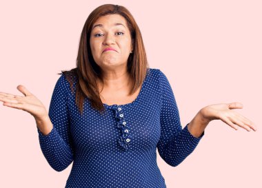 Orta yaşlı, gündelik kıyafetler giyen, elleri ve kolları havada, kafası karışmış bir Latin kadın. şüphe kavramı. 