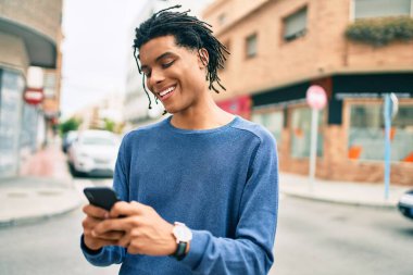 Genç Afrikalı Amerikalı adam şehrin caddesinde akıllı telefon kullanarak mutlu gülümsüyor..