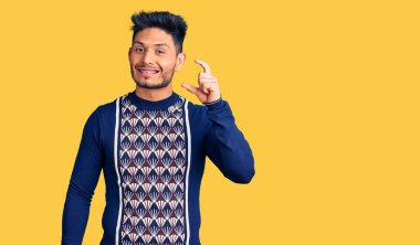 Yakışıklı Latin Amerikalı genç adam günlük kışlık süveter giyiyor gülümsüyor ve kendine güvenen el hareketleriyle küçük boy işareti yapıyor parmaklarıyla ve kamerayla bakıyor. ölçüm kavramı. 