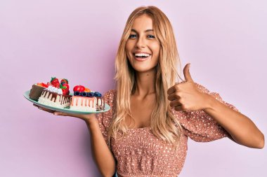 Güzel sarışın, elinde pasta dilimleri olan mutlu ve pozitif gülümseyen genç bir kadın. Başparmağın mükemmel ve onay işareti yapıyor. 