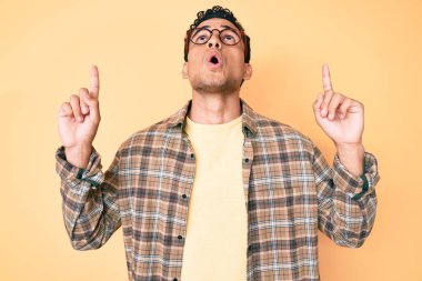 Genç, yakışıklı, günlük kıyafetler giyen, gözlüklü, şaşkın ve şaşırmış bir şekilde yukarı bakıp, parmaklarıyla işaret eden ve kollarını kaldıran bir İspanyol.. 