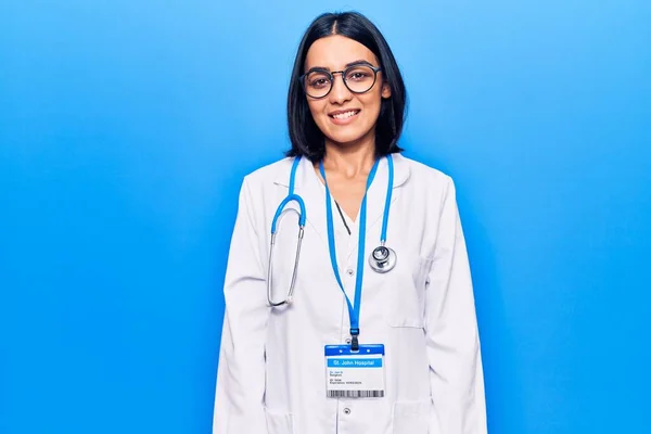 Young beautiful latin woman wearing doctor stethoscope and id card looking positive and happy standing and smiling with a confident smile showing teeth