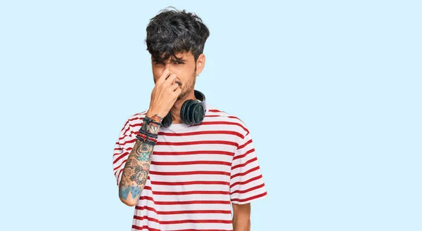 若いヒスパニック系の男性は ヘッドフォンを使用して音楽を聞いて何か臭くて嫌な 耐え難い匂いを嗅ぎ 鼻の上に指で息を保持します 臭いが悪い — ストック写真