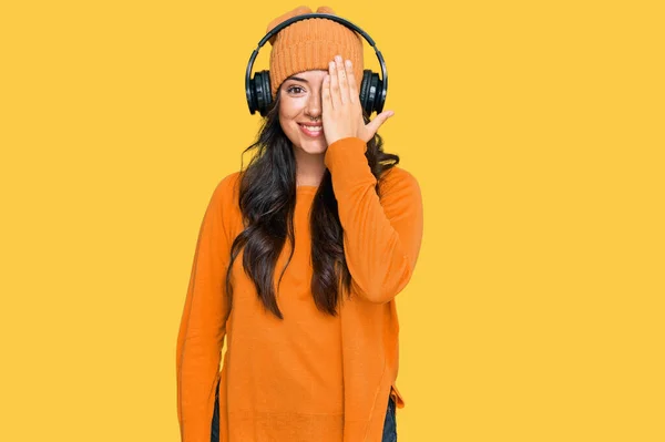 美しいブルネットの若い女性は 手で一つの目をカバーヘッドフォンを使用して音楽を聞いて 顔に自信を持って笑顔と驚きの感情 — ストック写真
