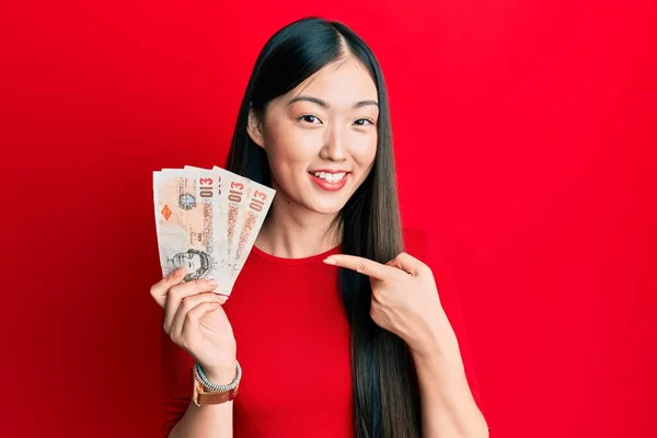 Young chinese woman holding 10 united kingdom pounds banknotes smiling happy pointing with hand and finger