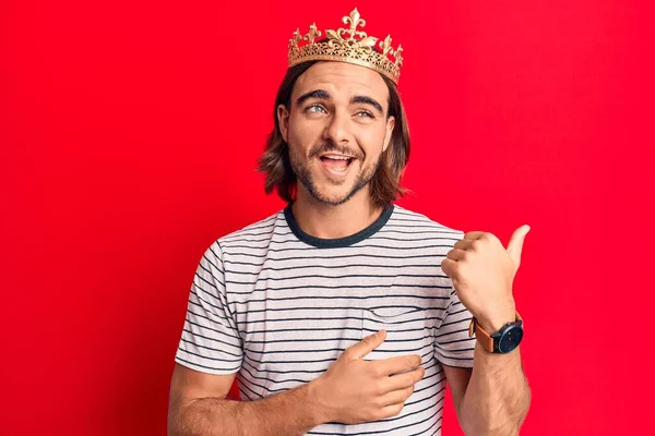 Young handsome man wearing prince crown pointing thumb up to the side smiling happy with open mouth