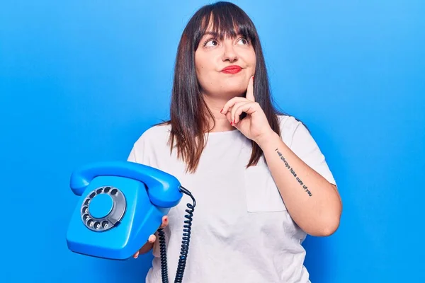 Young plus size woman holding vintage telephone serious face thinking about question with hand on chin, thoughtful about confusing idea