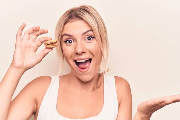 年轻的金发女人吃着法国甜点 白色背景下拿着巧克力玛卡伦 带着快乐的笑容和高傲的表情庆祝胜利 — 图库照片