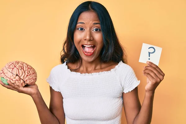 Young indian girl holding brain and question mark reminder smiling and laughing hard out loud because funny crazy joke.