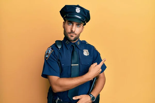 Handsome hispanic man wearing police uniform pointing with hand finger to the side showing advertisement, serious and calm face