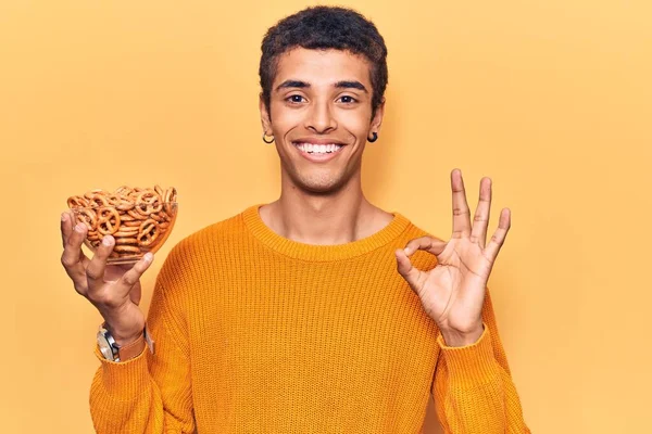 Young African Amercian Man Holding Pretzels Doing Sign Fingers Smiling — Stock Photo, Image