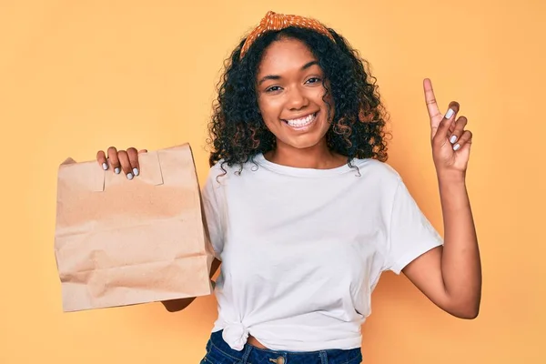 アフリカ系アメリカ人の若い女性は 幸せな顔をしたアイデアや質問を指差す指で驚いて紙袋を奪う ナンバーワン — ストック写真
