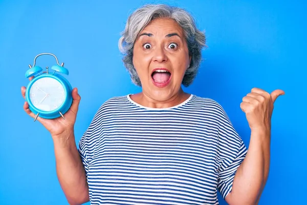 Senior Hispânico Mulher Segurando Despertador Apontando Polegar Até Lado Sorrindo — Fotografia de Stock