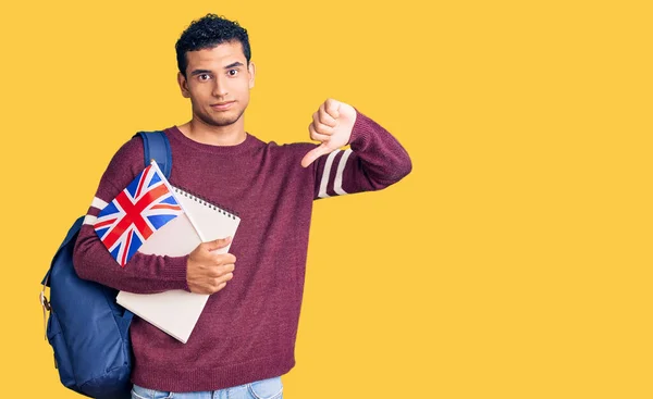 Hispanic handsome young man exchange student holding uk flag with angry face, negative sign showing dislike with thumbs down, rejection concept