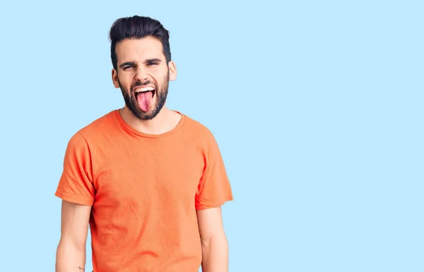 ひげを生やした若いハンサムな男が面白い表情で幸せ舌を突き出してカジュアルなTシャツを着ています 感情の概念 — ストック写真
