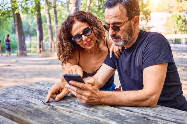 Medelålders Par Ler Glada Med Hjälp Smartphone Sitter Bänken Parken — Stockfoto