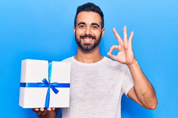 Молодий Іспаномовний Чоловік Тримає Подарунок Роблячи Знак Пальцями Посміхаючись Дружній — стокове фото