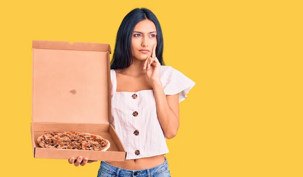 年轻美丽的拉丁女孩拿着披萨盒 严肃的面容 手托着下巴思考着问题 沉思着困惑的想法 — 图库照片