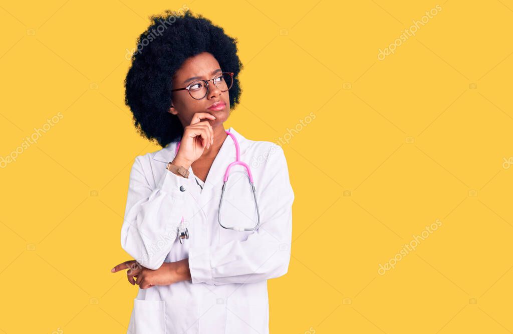 Young african american woman wearing doctor coat and stethoscope with hand on chin thinking about question, pensive expression. smiling with thoughtful face. doubt concept. 