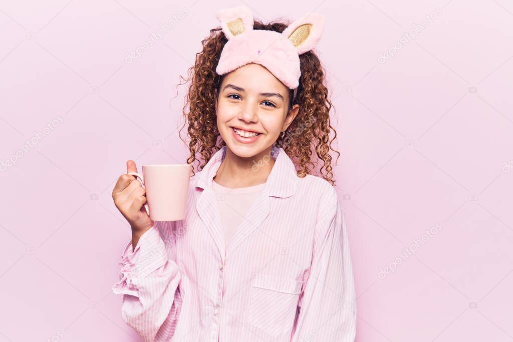 Beautiful kid girl with curly hair wearing sleep mask and pajamas holding coffee looking positive and happy standing and smiling with a confident smile showing teeth 