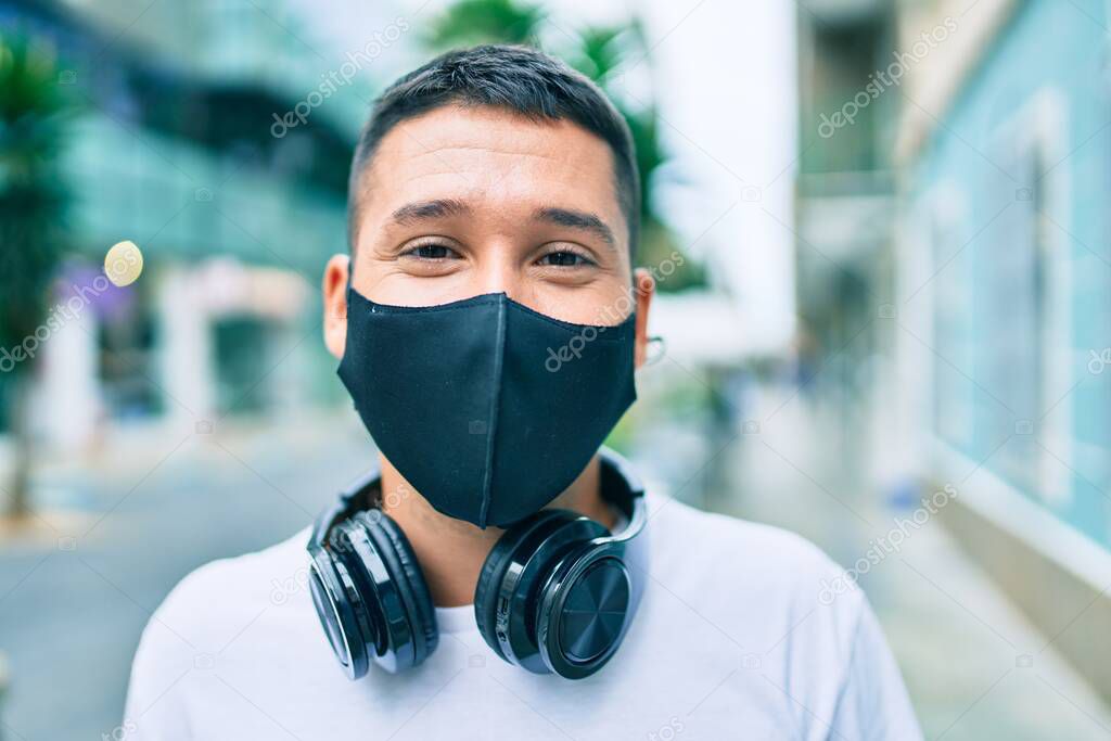 Young hispanic man wearing coronavirus protection mask using headphones at street of city.
