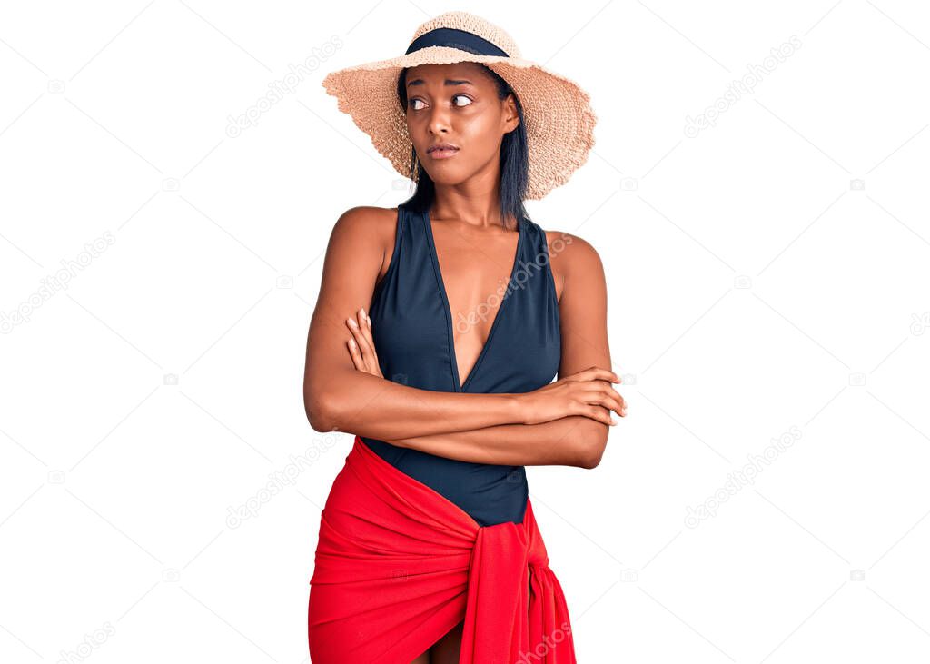 Young african american woman wearing swimsuit and summer hat looking to the side with arms crossed convinced and confident 
