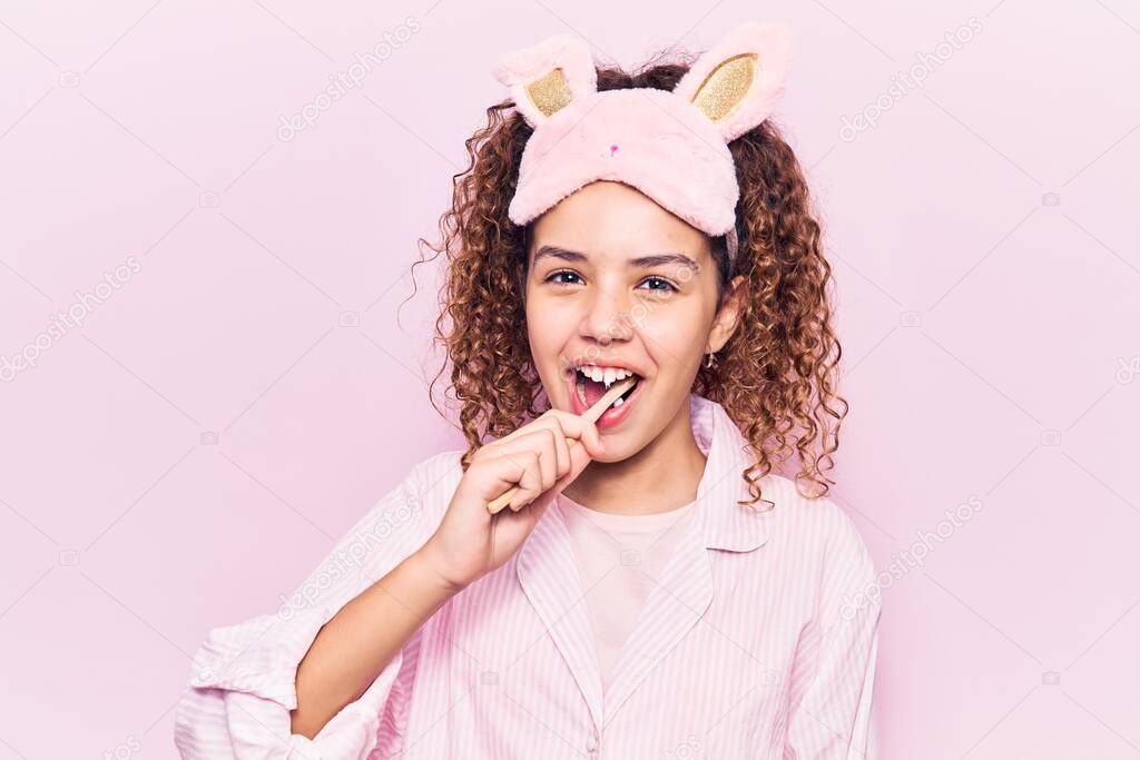 Beautiful latin teenager wearing pajama and sleep mask smiling happy. Standing with smile on face using toothbrush over isolated pink background