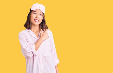 Genç, güzel Çinli kız uyku maskesi takıyor ve pijama giyiyor. Yüzünde mutlu ve doğal bir ifadeyle, elleri ve parmaklarıyla yüzünü gösteren bir gülümseme var. 