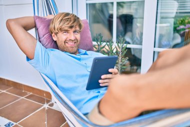 Orta yaşlı yakışıklı bir adam evinin terasında, bir hamakta dokunmatik bir aletle dinleniyor.