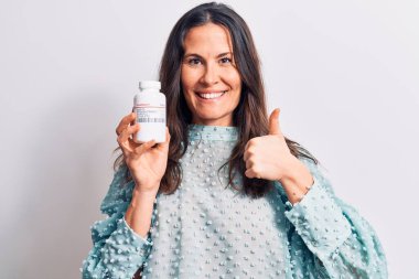 Genç, güzel, esmer bir kadın izole edilmiş beyaz arka planda ilaç dolu bir kavanoz tutuyor mutlu ve pozitif gülümsüyor, başparmağıyla mükemmel ve onay işareti yapıyor.