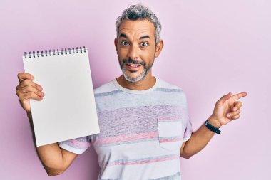 Orta yaşlı, gri saçlı, elinde boş bir defter tutan, gülümseyen, mutlu, eli ve parmağı yanda olan bir adam. 