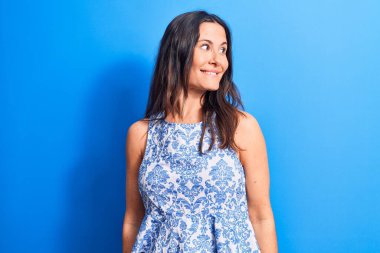 Genç, güzel, esmer bir kadın mavi arka plan üzerine kolsuz tişört giyiyor. Yan tarafa bakıyor, rahat bir poz veriyor ve kendine güvenen bir gülümseme sergiliyor..