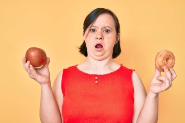 Down sendromlu esmer bir kadın kırmızı elma ve çörek tutuyor. Şok olmuş bir halde oturuyor. Şüpheci ve alaycı bakıyor. 