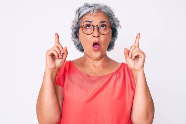 Son sınıf Latin kökenli, gri saçlı, günlük kıyafetler giyen, gözlüklü, şaşkın, şaşkın, parmaklarıyla işaret eden ve kollarını kaldıran bir kadın.. 