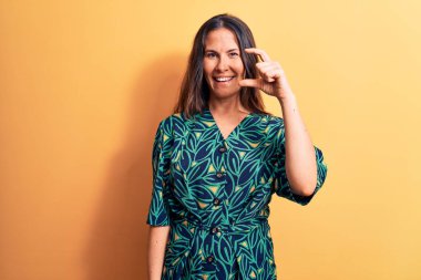 Genç, güzel, esmer, sıradan bir elbise giyen, sarı arka planda duran, gülümseyen, kendine güvenen, el hareketi yapan, küçük beden işareti yapan, parmaklarını gösterip kameraya bakan bir kadın. Ölçü kavramı.