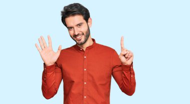 Latin asıllı genç adam gündelik giysiler giyip altı numaralı parmaklarıyla işaret ederken kendinden emin ve mutlu bir şekilde gülümsüyor.. 