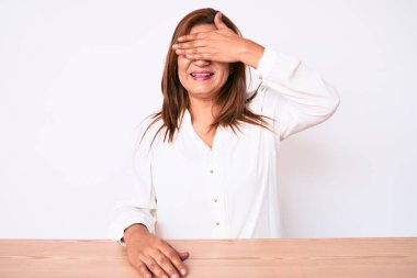 Orta yaşlı esmer, İspanyol iş kadını. Masada oturan, gülümseyen ve gülümseyen, yüzünde şaşkınlık için gözlerini kapatan sıradan bir beyaz gömlek giyiyor. kör kavram. 