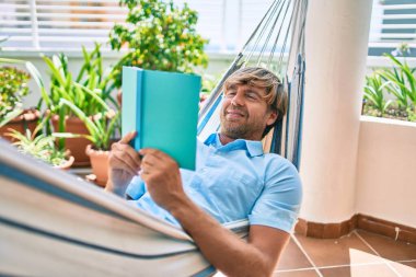 Orta yaşlı yakışıklı bir adam evinin terasında bir hamakta uzanıp kitap okuyor.