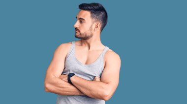 Mayo ve kolsuz tişört giyen genç yakışıklı adam kollarını kavuşturmuş ve kendinden emin bir şekilde yan tarafa bakıyor. 