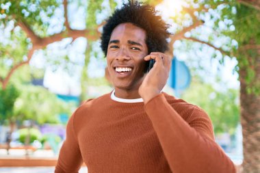 Genç, yakışıklı, Afro-Amerikan bir adam günlük giysiler giyip mutlu gülümsüyor. Yüzünde tebessümle dikilip şehrin caddesinde akıllı telefondan konuşuyor..