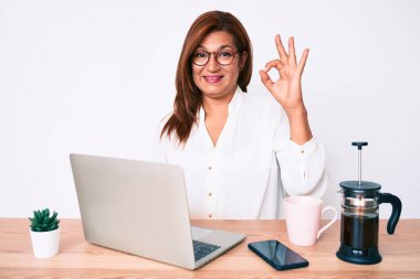 Orta yaşlı esmer, ispanyol iş kadını ofiste çalışıyor parmaklarıyla imza atıyor, gülümsüyor ve mükemmel bir sembol oluşturuyor. 