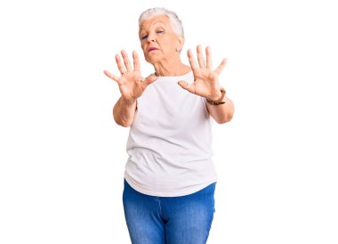 Mavi gözlü, gri saçlı, beyaz tişörtlü, el ve parmaklarıyla çerçeve yapan son sınıf güzeli kadın. 