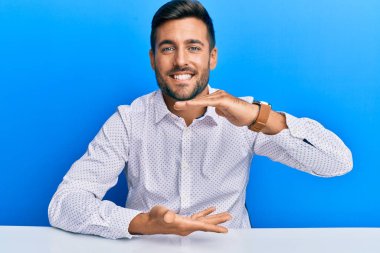 İş kıyafetleri giymiş yakışıklı İspanyol adam masaya oturmuş büyük ve büyük boy işareti gösteren el hareketleriyle ölçü sembolü gösteriyordu. Kameraya bakıp gülümsüyor. ölçüm kavramı. 