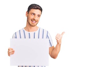 Elinde boş pankart tutan yakışıklı genç adam başparmağını yana kaldırmış, ağzı açık gülümsüyor. 