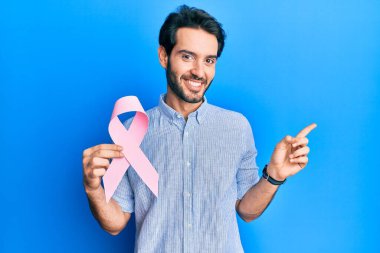 Genç İspanyol adam pembe kanser kurdelesi tutuyor gülümsüyor mutlu bir şekilde elini ve parmağını yana uzatıyordu. 