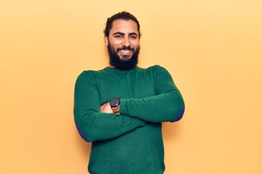 Genç Arap adam sıradan kıyafetler giyip kollarını kavuşturup kameraya bakarken gülümsüyor. pozitif kişi. 